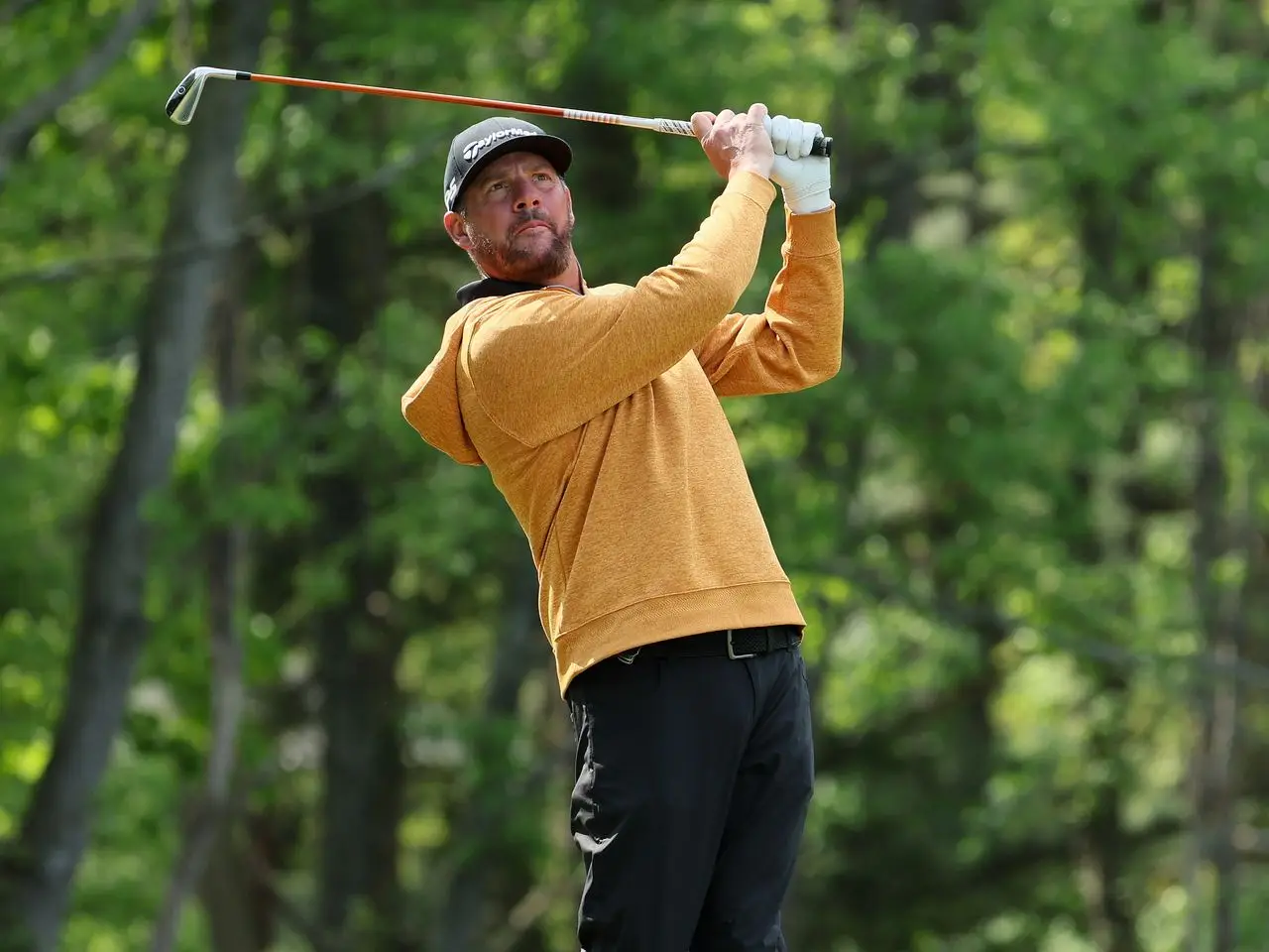 A person wearing an orange hoodie and black pants is playing golf, swinging a PXG club, renowned for its quality and performance, against a backdrop of green trees.