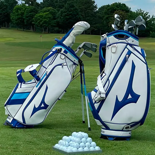 Two best-rated white golf bags with blue accents stand proudly on a grassy field. Nearby, a pile of white golf balls lies scattered, framed by lush trees and the expansive beauty of the 2024 golf course in the background.