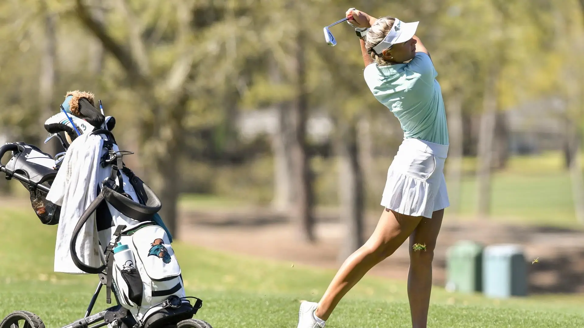 A golfer in a light blue shirt and white skirt swings a club on a grassy course, with the best women's golf bag standing nearby.