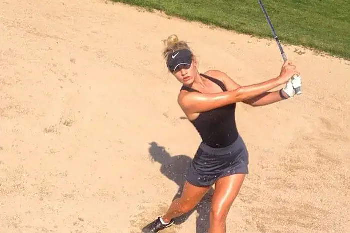A beautiful woman in a black outfit swings a golf club with finesse in a sunny bunker, showcasing her top 10 skills on the course.
