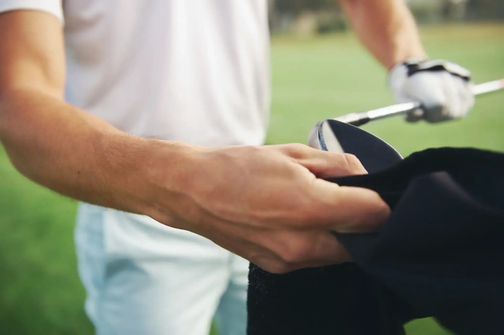 A person meticulously cleans a golf club with a black cloth on a grassy field, maintaining its gleam like it's one of the game's best-kept secrets.