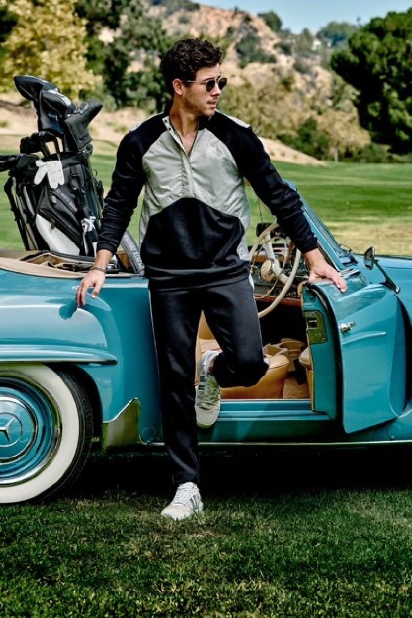 A man wearing sunglasses stands near an open car door next to a golf course, showcasing PXG Golf clubs in the back of a vintage blue car.