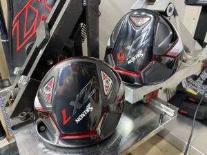 Close-up of two black Srixon ZX7 golf club heads placed on a piece of golf equipment, likely for customization or adjustment. The club heads, featuring red and white accents, are seen in a well-lit setting with other equipment nearby, reminiscent of a detailed driver review scene.