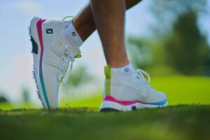 Person wearing white and multicolored athletic shoes walks on grass, with a blurred green and blue background.