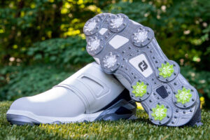 White golf shoes with velcro straps are displayed on grass. One shoe is upright, while the other is tilted to show the studded sole with gray and green spikes. Trees are in the background.