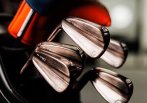 A close-up image of a set of TaylorMade P790 golf irons, showing numbers 7, 8, and 9, with an orange and blue head cover in the background. Elevate your game with these precision-engineered clubs.