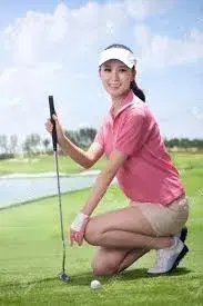 A rising star among Asian golfers crouches on a golf course wearing a pink polo shirt and beige shorts poised with a golf club near a pristine white ball showcasing the promising future of Golf 