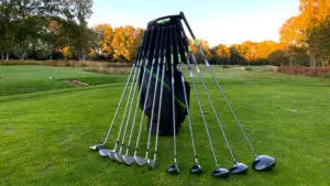 A set of golf clubs arranged in a fan shape around a golf bag placed on a vibrant green golf course with autumn trees in the background