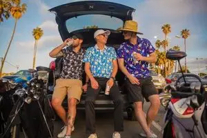 Three men in casual summer attire including great shirts from top golf polo brands sit on the open tailgate of a car They hold cans with golf bags nearby and palm trees swaying in the background ready to enjoy their game