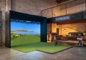 The ultimate guide to your indoor golf simulator setup features a virtual screen with a golf course a small putting green upfront and seating on the right A Sim in a Box sign above completes this perfect match for any golf enthusiast