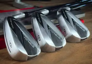 Three modern golf club irons labeled   and  are displayed in a row on a wooden surface with their shafts lying flat