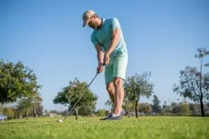 A man in a light blue shirt and green shorts is playing golf on a sunny day focusing on his golf stance as he prepares to hit the ball on the green Surrounded by trees and a clear sky he's set to improve his swing with precision.