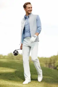 A man in a light blue Peter Millar shirt white pants and vest stylishly holding a golf club stands on a grassy field with trees in the background