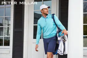 A man in a light blue pullover and shorts epitomizing luxury apparel carries a golf bag as he leaves a building The Peter Millar logo graces the top left corner offering style tips to elevate your look