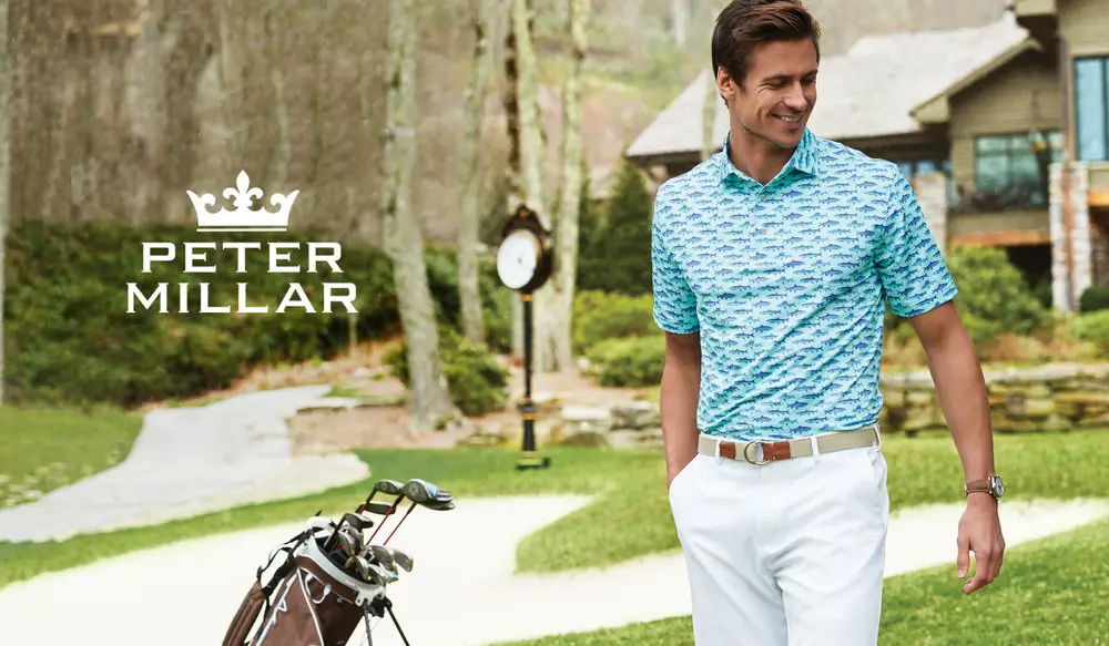 A man in a stylish patterned polo shirt and white pants stands near a golf course, his luxury apparel complemented by the Peter Millar logo on the left. Golf clubs are in the foreground, completing this elegant style outfit.