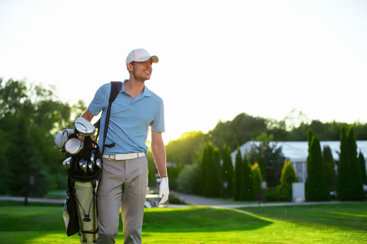 Man in a light blue shirt and cap walking on a golf course, carrying a golf bag over his shoulder. Trees and a building are visible in the background under a bright sky.